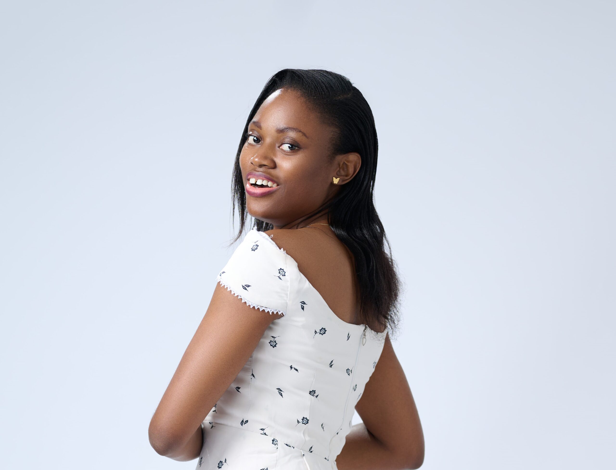 Portrait of Jane OLUWADARE, turned away from the camera, looking back over her shoulder with a bright smile. She is wearing a white dress and small gold butterfly earring studs. Her hair is down in a side part